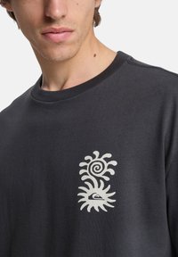 Σκούρο γκρι φούτερ με στρογγυλή λαιμόκοψη, απαλό ύφασμα και λευκό γραφικό σχέδιο με σπείρες και σχέδια ήλιου στην αριστερή περιοχή του στήθους.