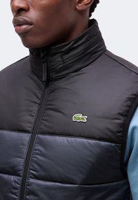 Gros plan d'une personne portant un gilet matelassé noir et bleu marine avec un logo crocodile vert sur la poitrine et une chemise bleu clair en dessous.