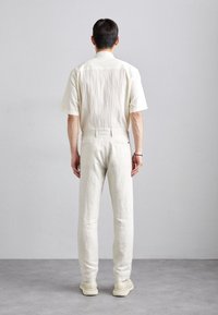 Kremowa koszula z krótkim rękawem z teksturowanej tkaniny, w parze z jasnymi lnianymi spodniami i beżowymi butami, widziana od tyłu.