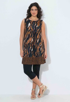 Mujer de pie contra una pared blanca, vestida con una túnica sin mangas con estampado, leggings negros recortados y sandalias de cuña doradas con tiras al tobillo.