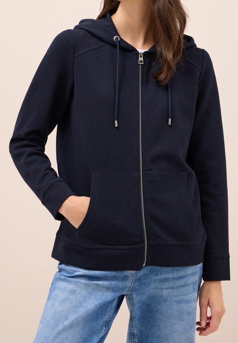 Femme portant un sweat à capuche bleu marine zippé avec poches avant et un jean bleu clair, debout avec une main dans la poche.