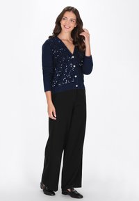 Cardigan navy con decorazioni in paillettes, scollatura a V, chiusura a bottoni e maniche lunghe, abbinato a pantaloni neri a gamba larga e ballerine.