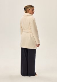 Kremowy szlafrok z polaru o teksturowanej powierzchni, z kołnierzem i paskiem w talii. Połączony z ciemnymi spodniami o szerokich nogawkach. Tło neutralne.