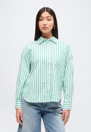 Žena s dlhými rovných vlasmi, ktorá má na sebe bielu košeľu so zelenými vertikálnymi pruhmi a svetlomodré džínsy, stojaca pred bielym pozadím.