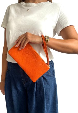 Clutch in pelle arancione con finitura testurizzata, chiusura con zip e laccio da polso, tenuta contro una camicia bianca e un pantalone in denim.
