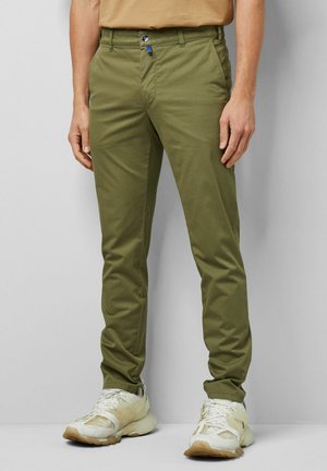 Mann trägt olivgrüne Slim-Fit-Hose und weiße Sneakers, steht vor einem einfarbigen hellgrauen Hintergrund.