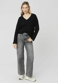 Pull noir à col en v avec coupe ample et manches longues associé à un jean gris taille haute et des baskets blanches, sur un fond neutre.