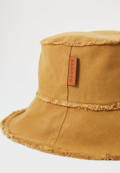 Kapelusz typu bucket w musztardowej żółci, z surowym brzegiem, warstwowym designem oraz skórzaną metką z wytłoczonym logo.