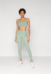 Grønn idretts-BH og matchende høyhårede leggings i et glatt, elastisk stoff, med subtile logo-detaljer. Fottøyet er hvite joggesko.