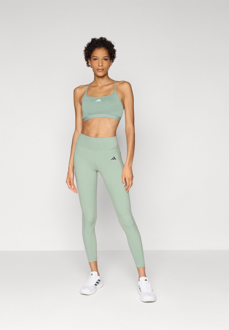 Reggiseno sportivo verde e leggings a vita alta abbinati in un tessuto liscio e elasticizzato, con dettagli del logo sottili. Le scarpe sono sneakers bianche.