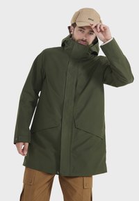 Grüne wasserdichte Jacke mit hohem Kragen, zwei großen Fronttaschen und sichtbaren Druckknöpfen. Getragen über einer beigen Mütze und braunen Hosen.