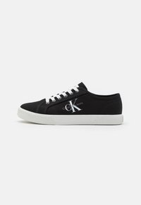 Svart Calvin Klein sneaker med vita snören, vit gummisula och "ck Calvin Klein Jeans"-logotyp på utsidan.