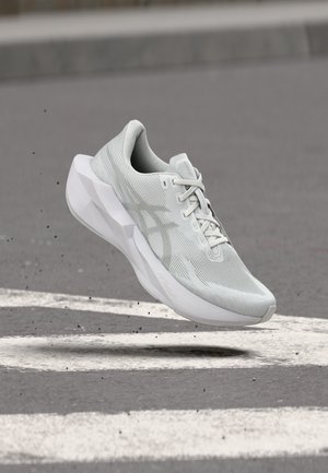 Lichtgrijze sportschoen met dikke zool lijkt boven asfalt met witte wegmarkeringen te zweven.