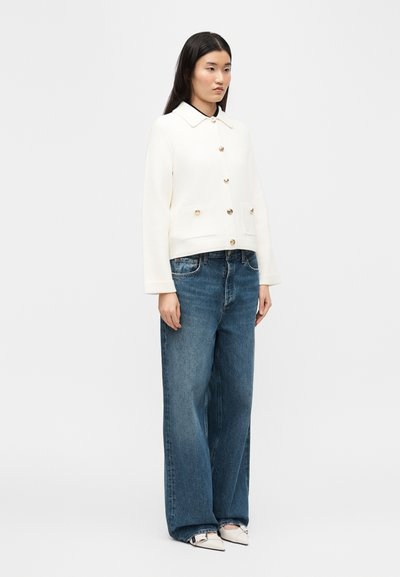 Γυναίκα με ίσια μαύρα μαλλιά στέκεται φορώντας λευκό σακάκι με κουμπιά, μπλε τζιν φαρδιάς γραμμής και μυτερές λευκές παπούτσια με μαύρες λεπτομέρειες.