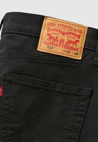 Zwarte denim broek met een achterzak met contrasterende stiknaden en een tan leren label met rode bedrukking, dat maat en merk aangeeft.