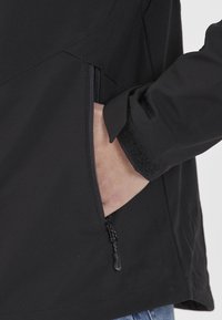 Prawa ręka wsunięta do zamykanej kieszeni czarnej kurtki z regulowanym mankietem na rzep, na białym tle.