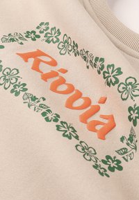 Tissu beige avec texte orange en relief "Ribbià" entouré d'une couronne florale verte près d'un col côtelé.