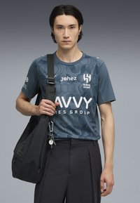 Jersey atlético de manga corta, azul, con patrones circulares y logotipos en blanco. Bolsa de hombro negra con llavero en la mano. Pantalones oscuros.