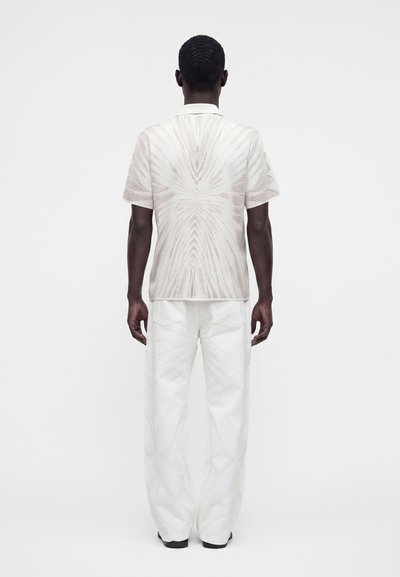 Homme debout, dos tourné, portant une chemise blanche à manches courtes à motifs et un pantalon blanc ample sur un fond blanc uni.