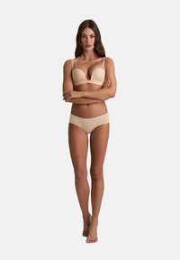 Donna in piedi con le braccia incrociate, indossa un reggiseno e delle mutande beige su uno sfondo grigio chiaro uniforme.