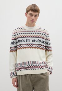 Cremefarbener Strickpullover mit einem geometrischen Muster in Rot, Navy und Gold, mit dem Schriftzug "APRÈS SKI" und Rippstrick-Akzenten an Bündchen und Saum.