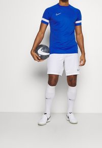 Hombre con camisa azul y pantalones cortos blancos sosteniendo un balón de fútbol blanco y negro, vistiendo calcetas y zapatos blancos con logotipos de Nike.