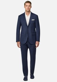 Costume navy fait en tissu texturé avec deux boutons, revers crantés et poches avant. Porté sur une chemise blanche, associé à des chaussures foncées.
