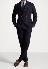 Tmavě modrý kostkovaný dvouřadý oblek s odpovídajícími kalhotami, světle modrá košile, vzorovaná kravata a hnědé mokasíny se zelenými ponožkami.