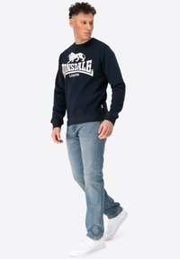 Navy crewneck sweatshirt with white lion logo, paired with light blue jeans and white sneakers. Slim fit, ribbed cuffs, and hem.