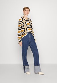 Blusa de manga longa com um fundo bege, padrões ovais em preto e amarelo. Combinada com jeans de cintura alta azuis com detalhes em denim.