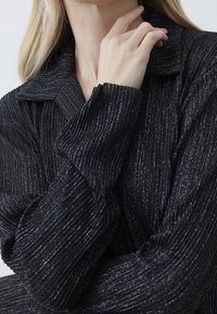 Černá lehká blůzka s texturovaným ribbovým designem a metalickými nitěmi, s límečkem a prodlouženými rukávy.