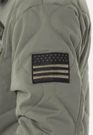 Patch de drapeau américain noir et gris cousu sur le haut du bras d'une veste matelassée vert olive.