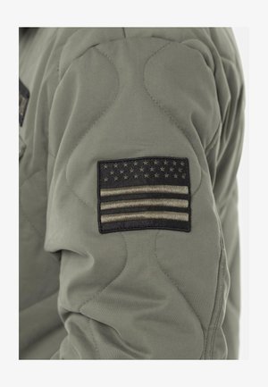 Patch de drapeau américain noir et gris cousu sur le haut du bras d'une veste matelassée vert olive.