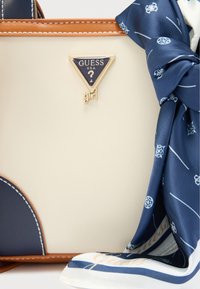 Τσάντα χειρός σε κρεμ χρώμα με σκούρες μπλε λεπτομέρειες, που διαθέτει τριγωνικό χρυσό λογότυπο «GUESS» και μαντήλι με μοτίβο στα σκούρα μπλε δεμένο στη λαβή. Καφέ δερμάτινες λεπτομέρειες.