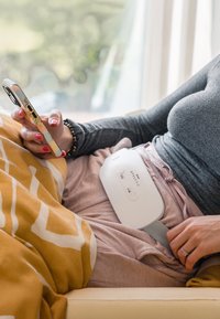 Weißes elektronisches Gerät mit Tasten, auf dem Schoß einer Person platziert, mit einem grauen Riemen gesichert. Weiche, strukturiert Decke im gelb-weißen Muster.