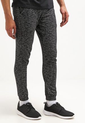 Le bas du corps d'une personne portant un pantalon de jogging gris foncé texturé, des baskets noires avec semelles blanches et des chaussettes blanches sur un fond uni.