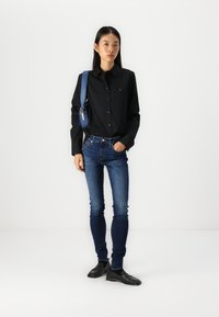 Zwarte button-up shirt, blauwe jeans en zwarte schoenen. Het shirt heeft een klein logo; de jeans zijn slim fit met een donkere wassingen. Inclusief blauwe tas.