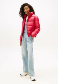 Red puffer jacket with a high collar and quilted design, worn over light blue denim overalls. Model stands in white background, wearing white sneakers.