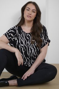 Černobílý top s krátkým rukávem se vzorem zebry, vyrobený z lehkého materiálu. Modelka sedí s překříženýma nohama a má na sobě černé kalhoty a boty.