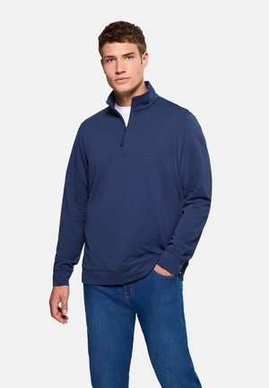 Joven vestido con un pulóver azul marino con cremallera hasta un cuarto, camiseta blanca debajo y vaqueros azules, de pie con una mano en el bolsillo.