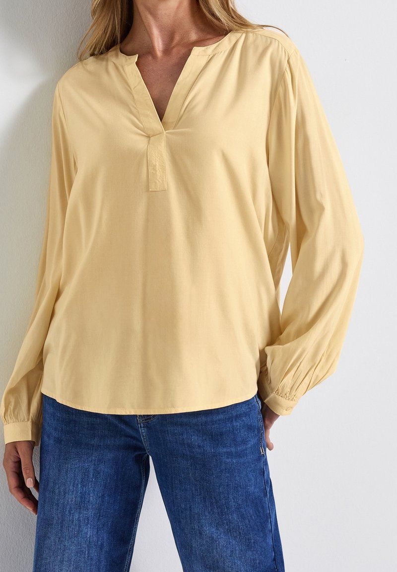 Femme portant un chemisier ample jaune pâle à manches longues avec un col en V et un jean bleu, debout contre un mur clair uni.