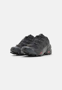 Chaussures de trail noires avec une tige texturée, semelles extérieures en caoutchouc solides et un système de laçage distinctif, présentant des accents rouges et un design robuste.