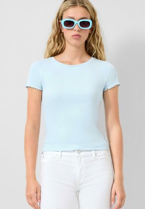 Junge Frau trägt ein hellblaues, geripptes Kurzarmshirt, weiße Hose und rechteckige Sonnenbrille in Himmelblau, steht vor einem schlichten, hellen Hintergrund.