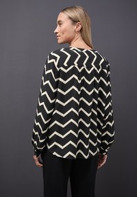 Schwarze Bluse mit cremefarbenem Zickzack-Muster, langen Ärmeln, lockerer Passform und geknöpften Manschetten. Rückenschlitz und Nackendetail sichtbar.