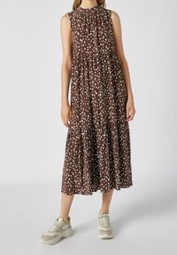 Brun, ärmlös maxiklänning med plisserad halsringning, som har ett vitt prickigt mönster och ett lager-på-lager-design, kombinerad med ljusa sneakers.