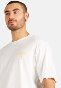 Hvid bomulds t-shirt med korte ærmer, der har et gult grafik, der læser "UNKNOWN" sammen med et solstråle design. Blød tekstur, afslappet pasform.
