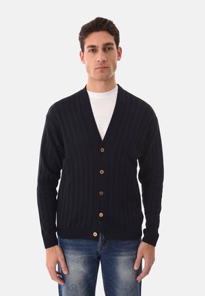 Uomo che indossa un cardigan nero con bottoni sopra una t-shirt bianca e jeans blu, in piedi davanti a uno sfondo chiaro e semplice.