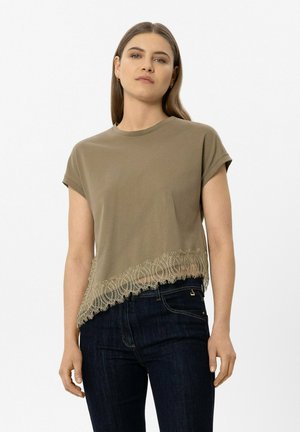 Woman with long brown hair wears an olive green short-sleeve top with lace trim and dark blue jeans, standing against a plain background.