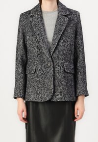 Blazer mit Fischgrätmuster in Schwarz und Weiß, mit Reverskragen, Knopfleiste vorne und zwei seitlichen Taschen. Getragen über einem grauen Oberteil.