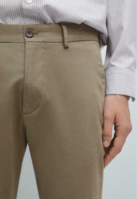 Khaki bukser med en glat tekstur, der har en knaplukning og sidelommer. Kombinationen er med en lysstribet skjorte.
